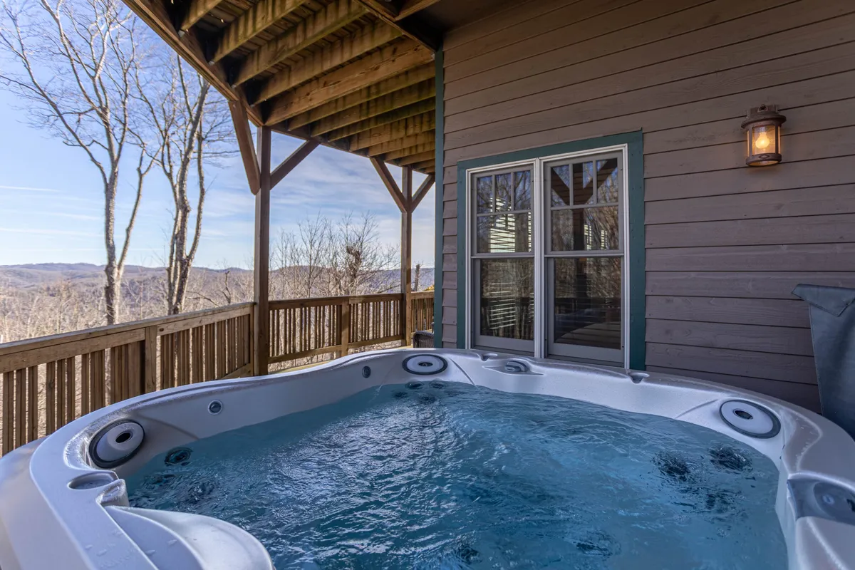 This image features an inviting outdoor hot tub set on a wooden deck with a stunning mountain backdrop. The scene includes a sturdy wooden railing and the exterior wall of a lodge-style building. The ambiance is tranquil, with hints of sunset lighting and a serene mountain landscape stretching beyond the deck.