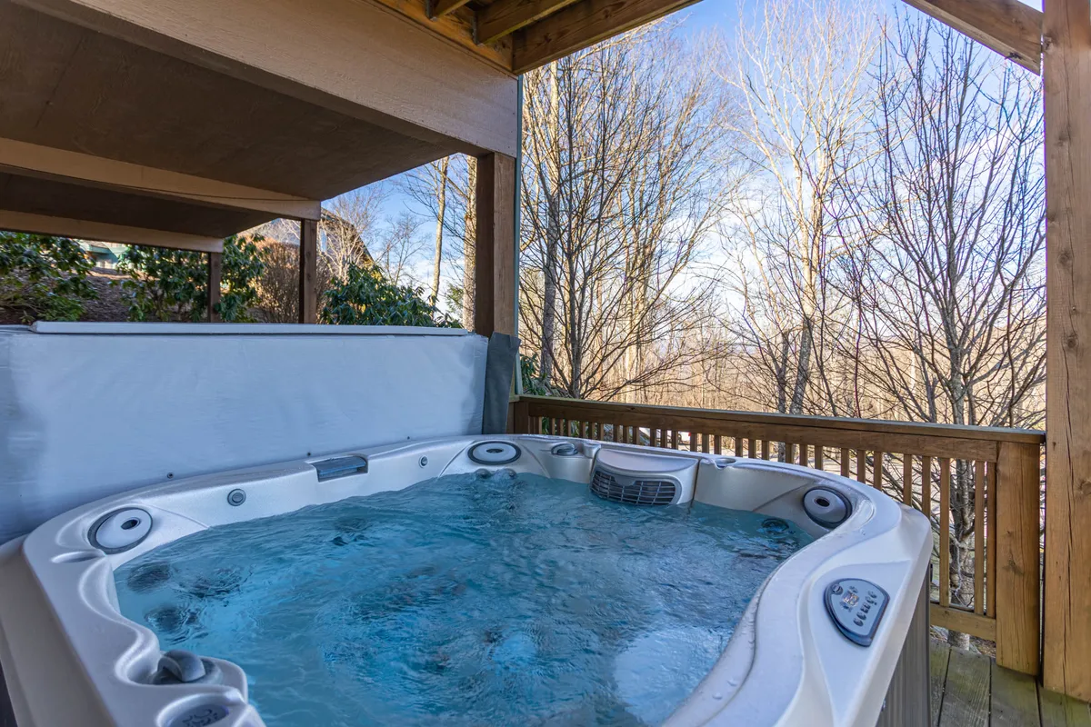 A wooden deck with an outdoor hot tub nestled among the trees, offering privacy and immersion in nature. The hot tub overlooks the surrounding forest, creating an ideal setting for relaxation and socializing under the open sky, whether by day or at sunset.