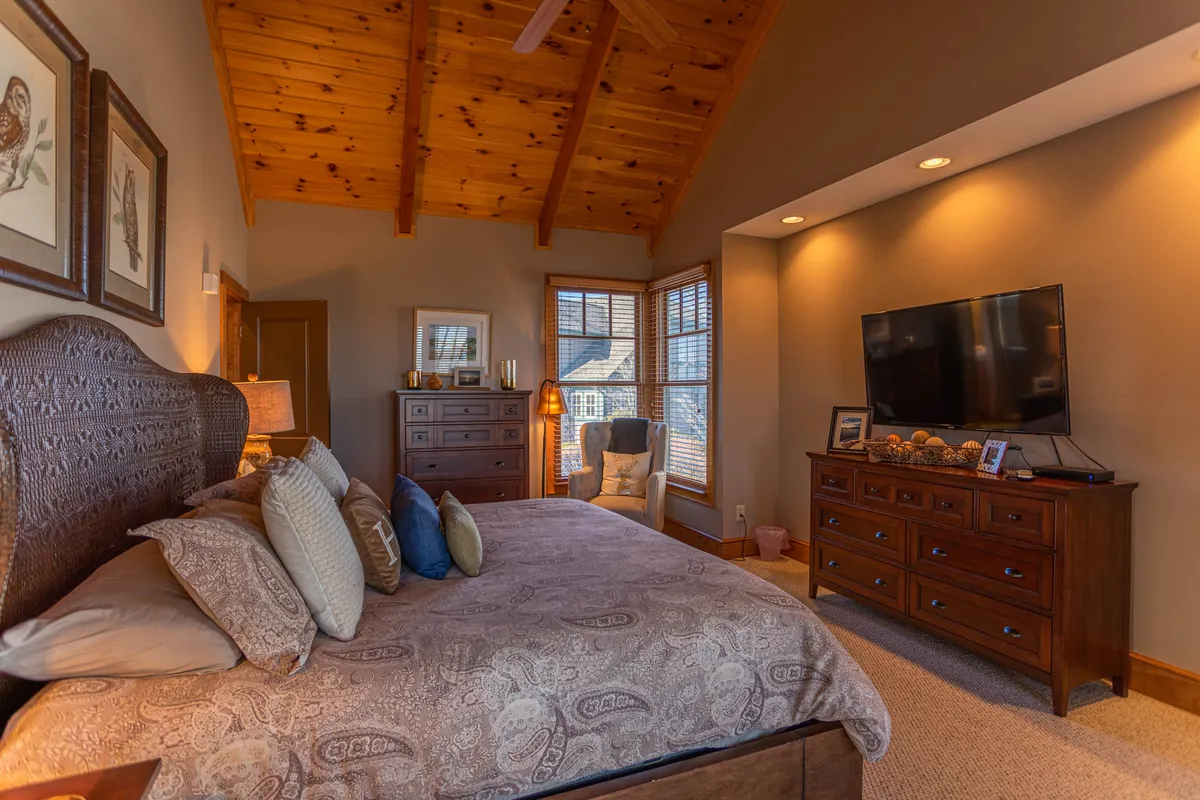 A spacious bedroom with a large wooden bed, mounted television, window that brings in natural light, a chest of drawers, and a reading chair. The mood is comfortable and serene, highlighting both practicality and relaxation.