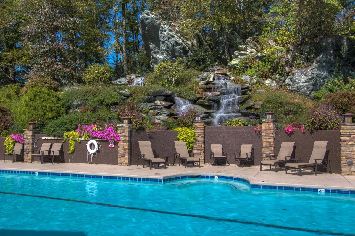 This exterior image showcases a sparkling swimming pool surrounded by lush landscaping, lounge chairs, and a cascading waterfall feature. The pool area is positioned to capture sunset lighting, with nature and comfortable seating inviting group gatherings and leisure activities.