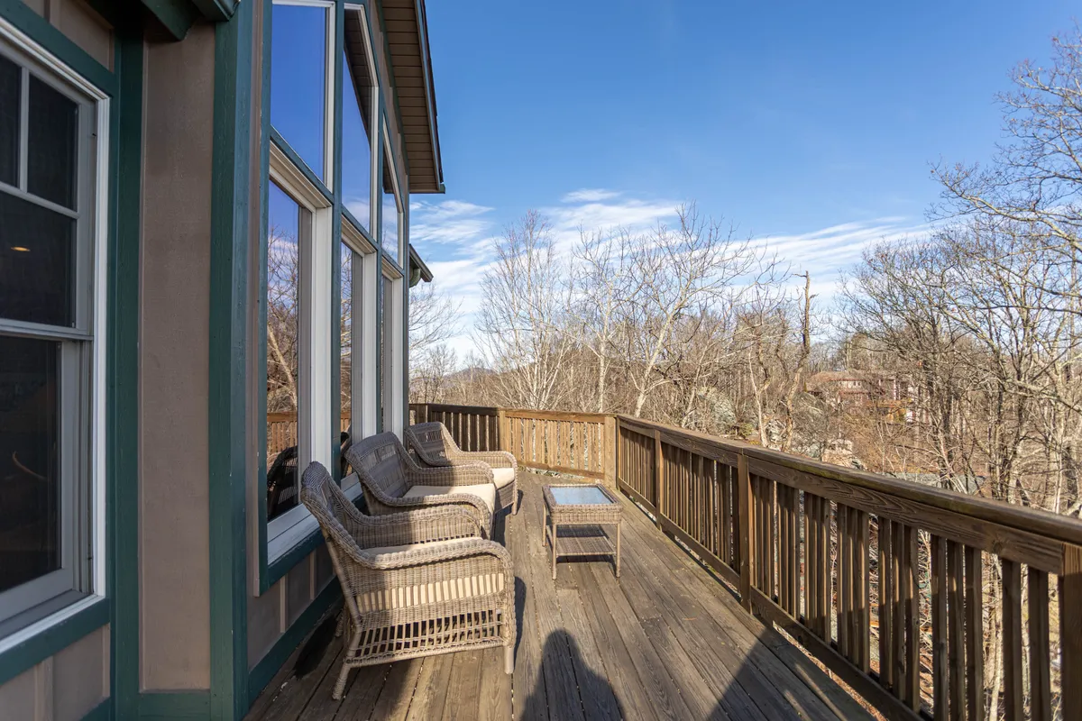 A peaceful deck set with wicker chairs and a glass table overlooks the trees beyond, enclosed by a wooden railing and complemented by large windows that invite the outdoors in. The space is ideal for quiet moments, morning coffee, or socializing after a day exploring.
