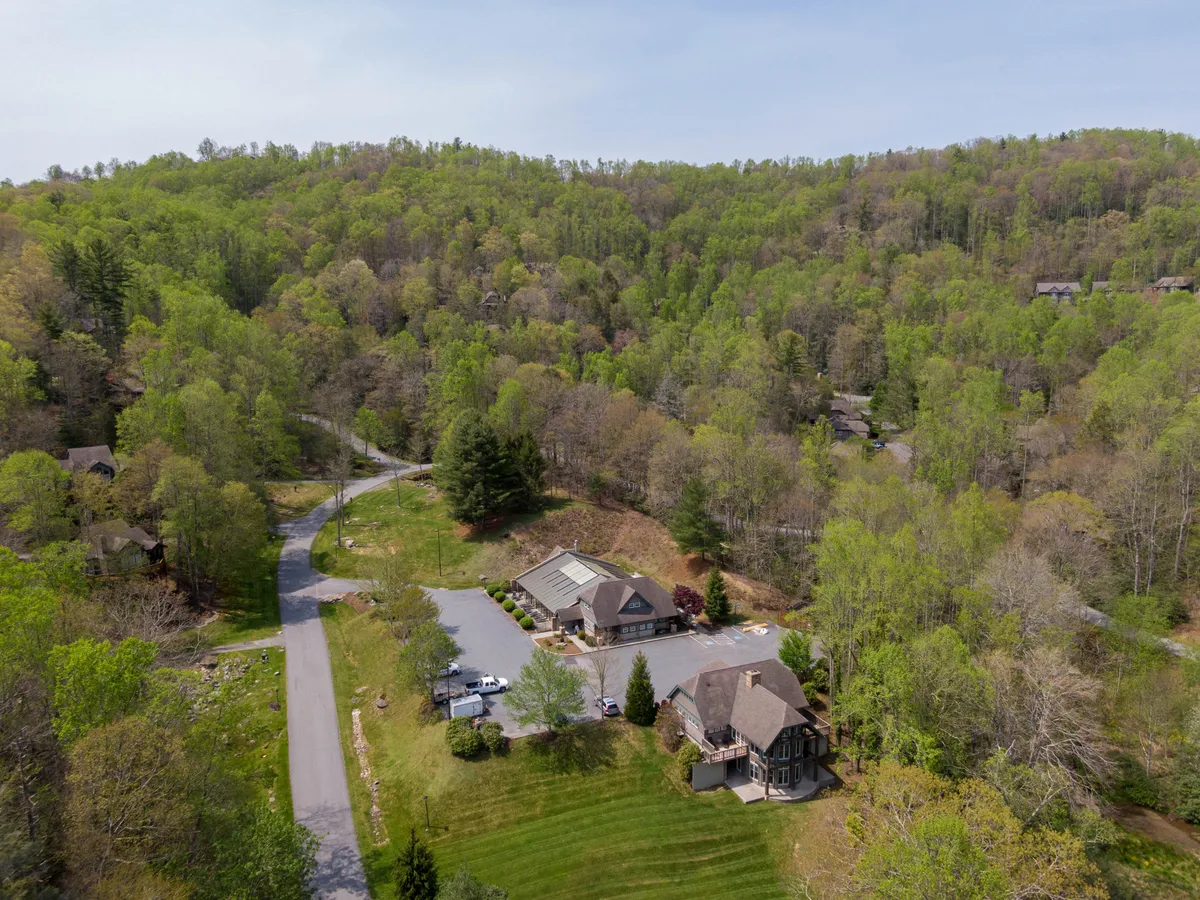 An exterior view of the main building framed by wooded hills, with a driveway, parking area, and walking paths leading into the landscape. The setting feels peaceful and inviting, capturing the larger context of the property within the mountain environment.