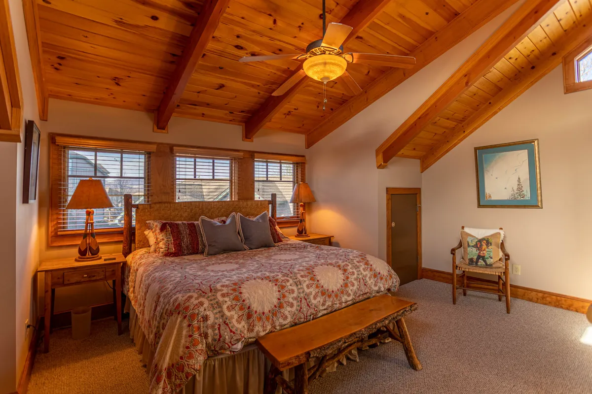 A master bedroom featuring a large bed with decorative bedding, wooden beams in the ceiling, and a window that fills the space with natural light. Nightstands with lamps offer functionality and the overall tone is warm and welcoming.