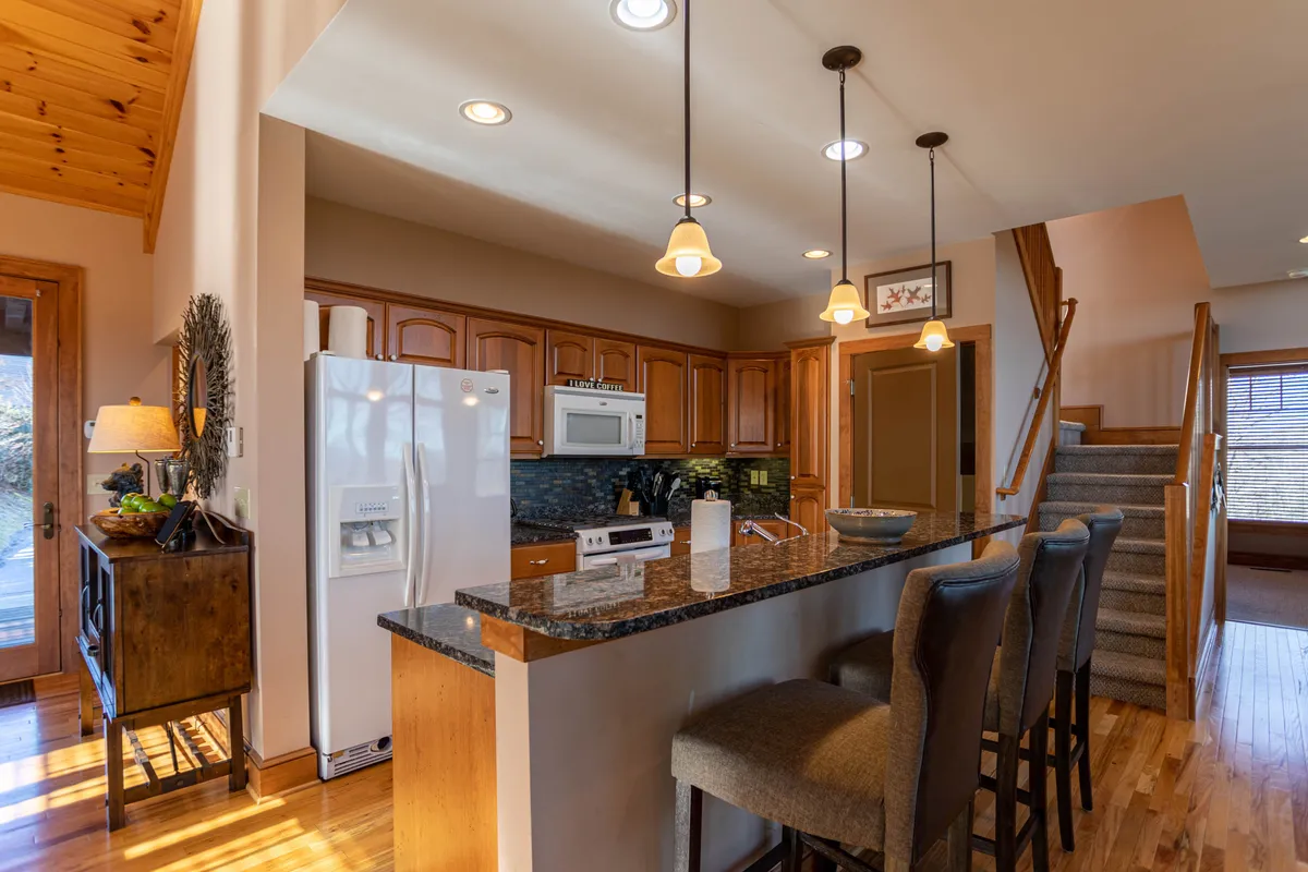 A gourmet kitchen with a granite countertop, kitchen island, stainless steel appliances, and wooden cabinetry. The scene is warm and welcoming, ideal for cooking, entertaining, and sharing meals. The staircase hints at the home's spacious layout and connectivity.