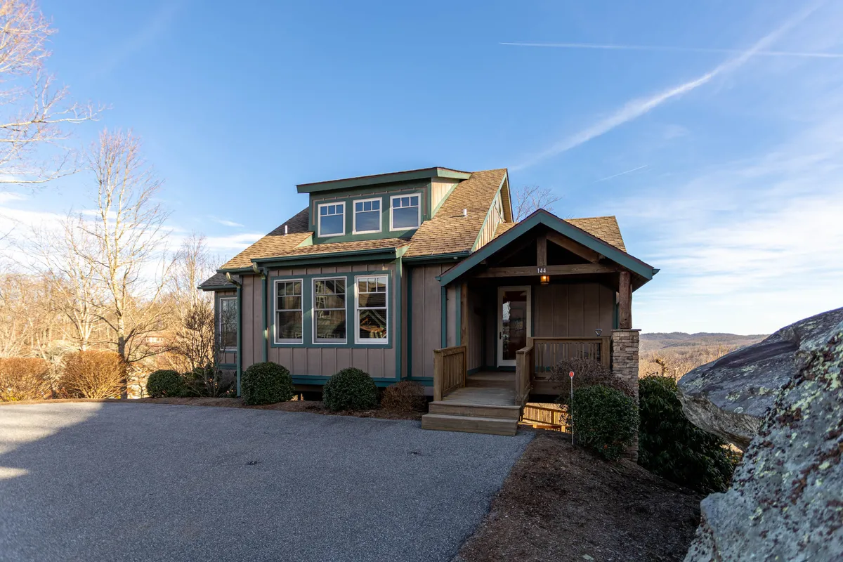 The exterior image showcases B Rock Ridge's inviting front porch, expansive windows that capture mountain views, distinctive rooflines, and beautifully landscaped gardens. Soft morning light streams across the porch, suggesting a perfect spot to relax and take in the tranquil surroundings, while the gardens reflect the seasonal charm of Banner Elk.