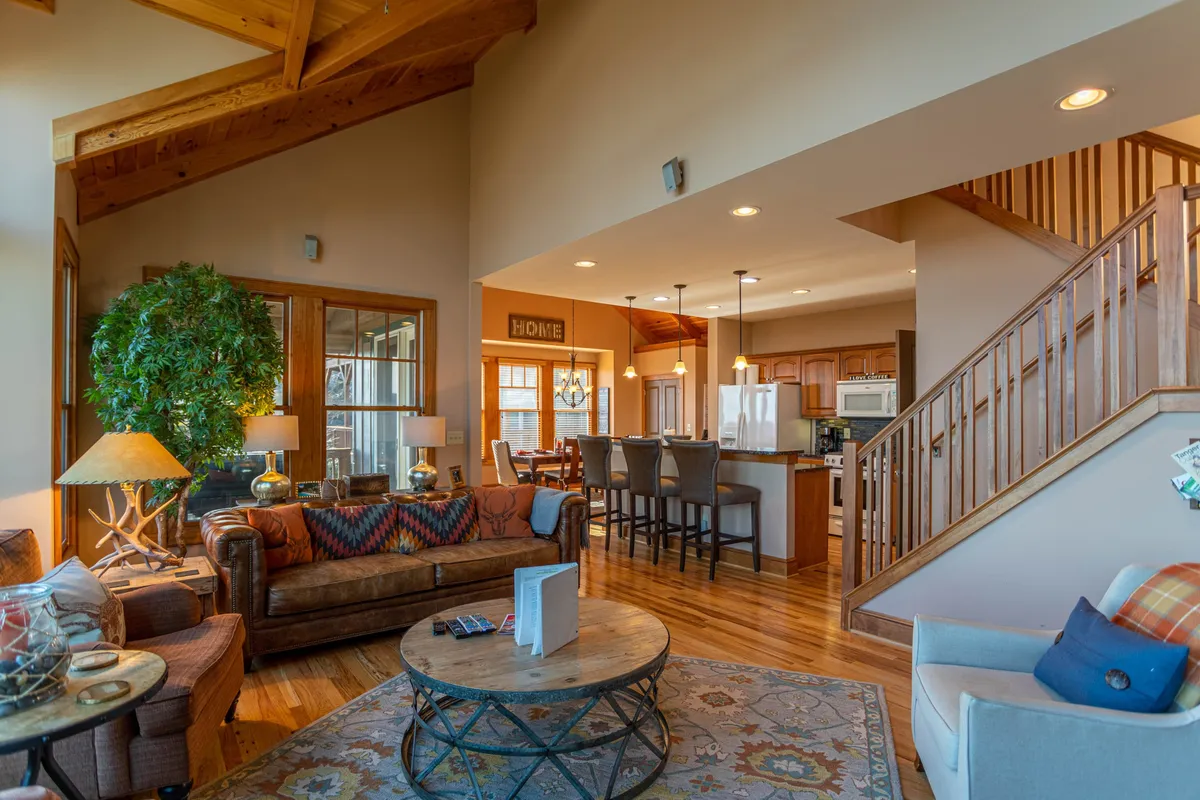 This image presents a spacious, open-concept living room with a large, comfortable sofa and a decorative coffee table in the foreground. The kitchen area is visible in the background, highlighting the seamless flow between the living and kitchen spaces. A staircase leads to the upper floor, enhancing the sense of openness.