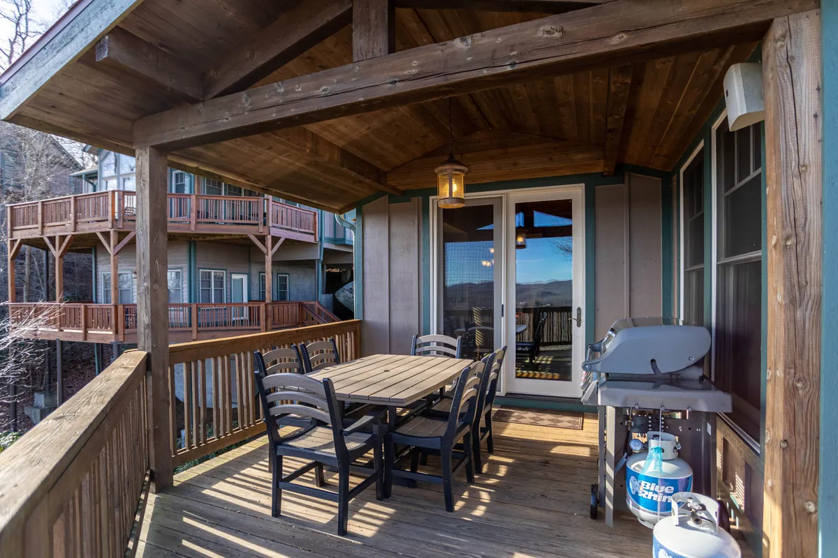 A welcoming deck scene featuring a wooden dining table surrounded by black chairs, a grill ready for barbecues, and a hanging lantern that adds warm ambiance. The deck is partially covered, with open air sections leading the eye to distant mountain views through the glass door, suggesting a seamless transition between indoor and outdoor living.