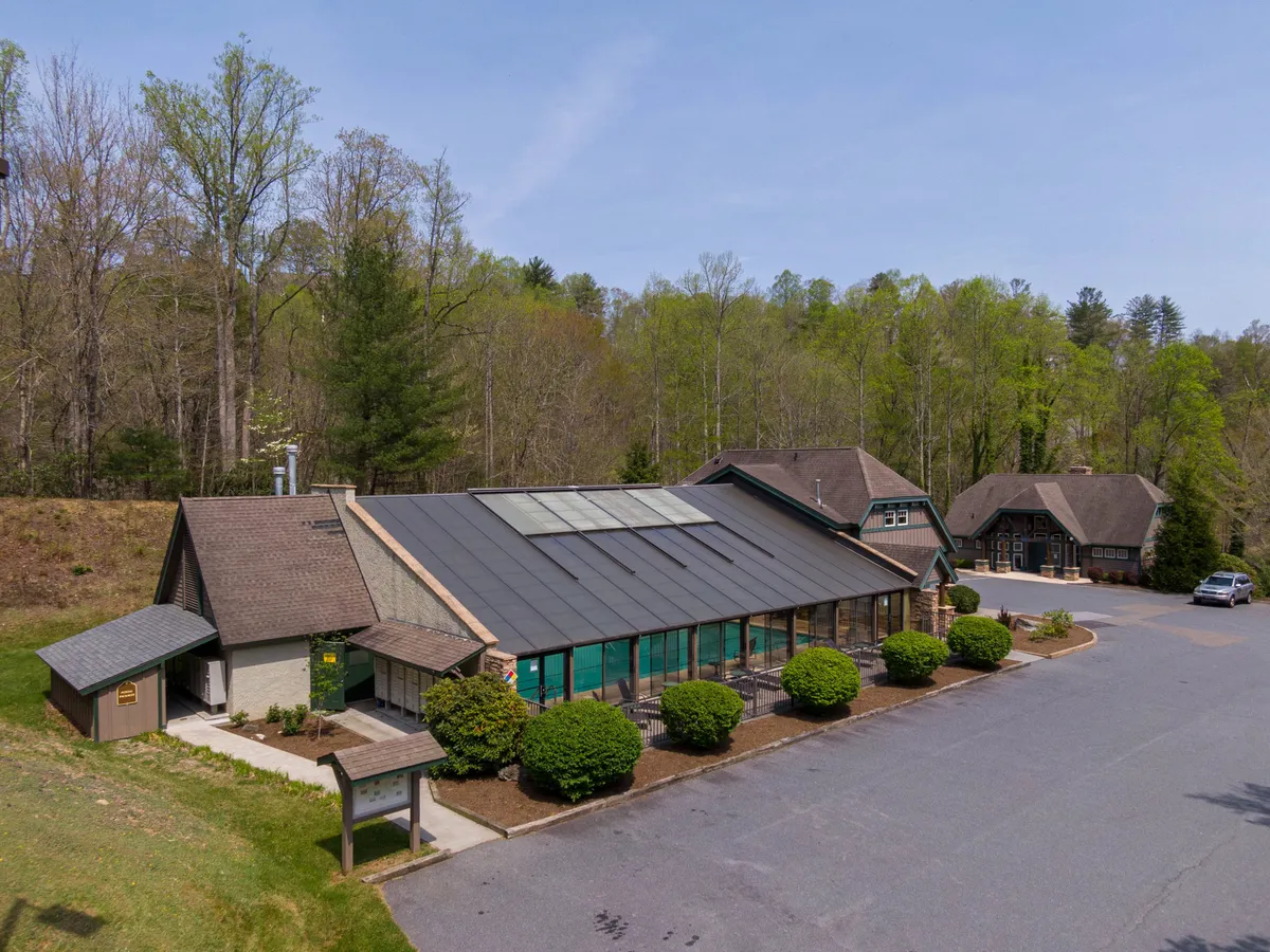 A warm, welcoming exterior shot of the main building with solar panels, surrounded by greenery and an adjacent smaller building. The parking area suggests convenience for group arrivals, and the lush landscape hints at communal spaces for events or relaxation. The scene radiates a sense of camaraderie and possibility for memorable group activities.