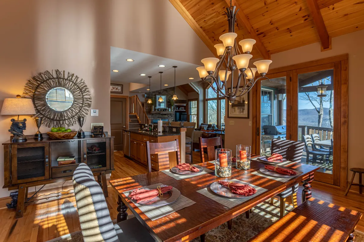 A beautifully set dining table with place settings, a striking chandelier above, and large windows that frame sweeping mountain views. Wooden beams and ample natural light add to the welcoming, cozy mood.